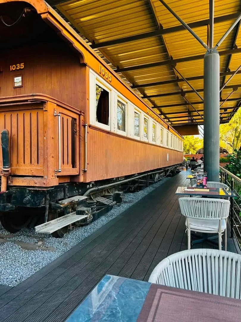 A historic railway carriage under the steel canopy, with the cafe's timber deck alongside
